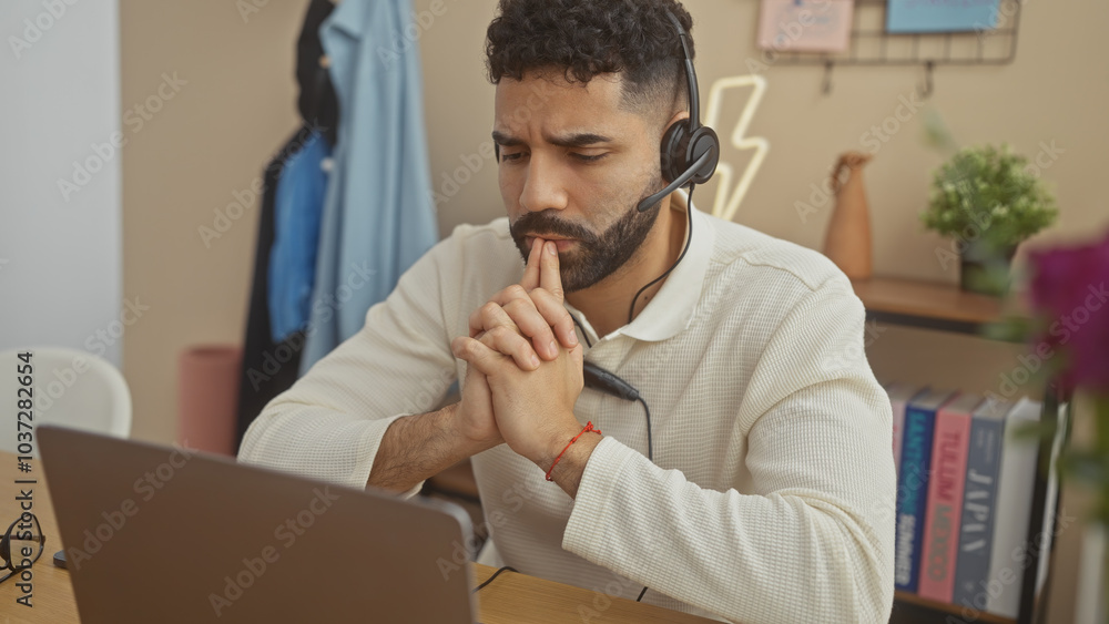 Fototapeta premium A thoughtful hispanic man with a beard wears a headset while working on a laptop in a cozy home office.