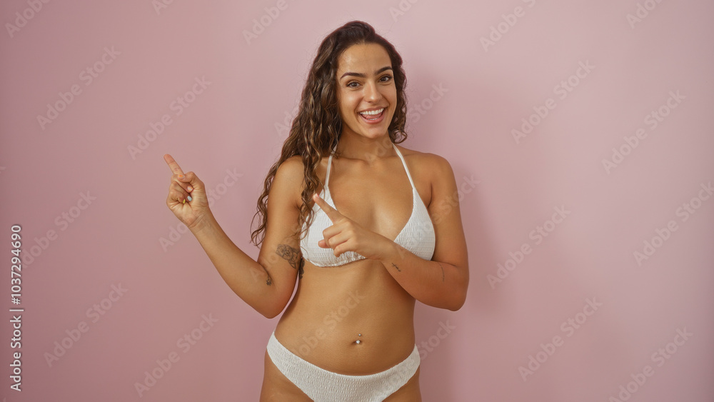 Naklejka premium Young woman pointing sideways against a pink wall, wearing a white bikini, smiling with a confident expression