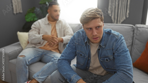 Men sitting in a living room, wearing casual clothes, with one looking away while the other talks, conveying a serious conversation taking place indoors in an apartment.