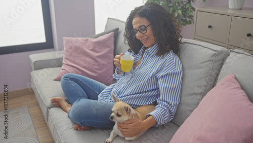 A middle-aged hispanic woman relaxes with her chihuahua on a sofa in a cozy living room, holding a mug.