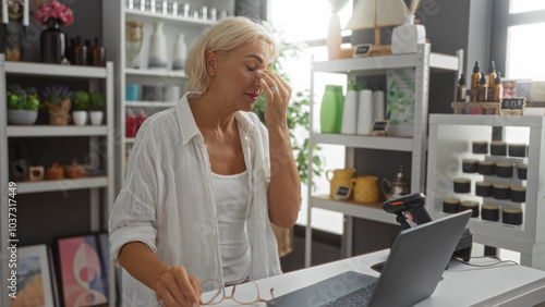 Woman working in a home decor shop looks stressed while sitting at a desk with a laptop, surrounded by various decorative items on shelves, creating a cozy and stylish indoor environment.