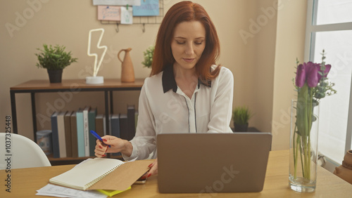 A focused woman with red hair writing notes beside a laptop in a well-organized home office