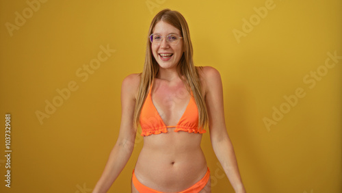 Young, attractive, blonde woman smiling cheerfully in an orange bikini against a vibrant yellow background wall, isolated and beautiful.