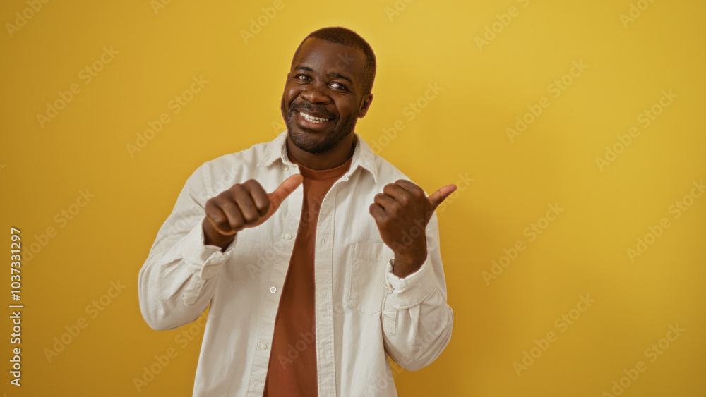 Young man smiling confidently pointing thumbs sideways against a vibrant yellow background, showcasing positivity and charisma in a casual yet impactful way. Young man smiling confidently pointing thumbs sideways against a vibrant yellow background, showcasing positivity and charisma in a casual yet impactful way.