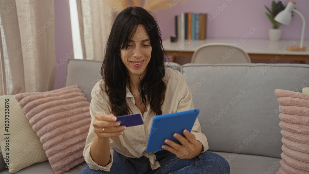 Woman smiling while shopping online using a tablet and credit card sitting on the sofa in a cozy living room with modern decor and natural light. Woman smiling while shopping online using a tablet and credit card sitting on the sofa in a cozy living room with modern decor and natural light.