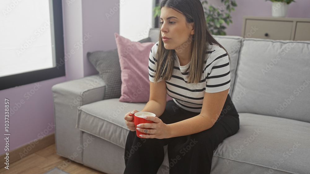 A reflective woman sits indoors, holding a mug, in a striped shirt, contemplating in a modern living room. A reflective woman sits indoors, holding a mug, in a striped shirt, contemplating in a modern living room.