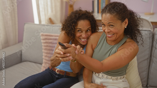 Two hispanic women laughing and playfully fighting over a tv remote in a cozy living room, showcasing familial love and joy.