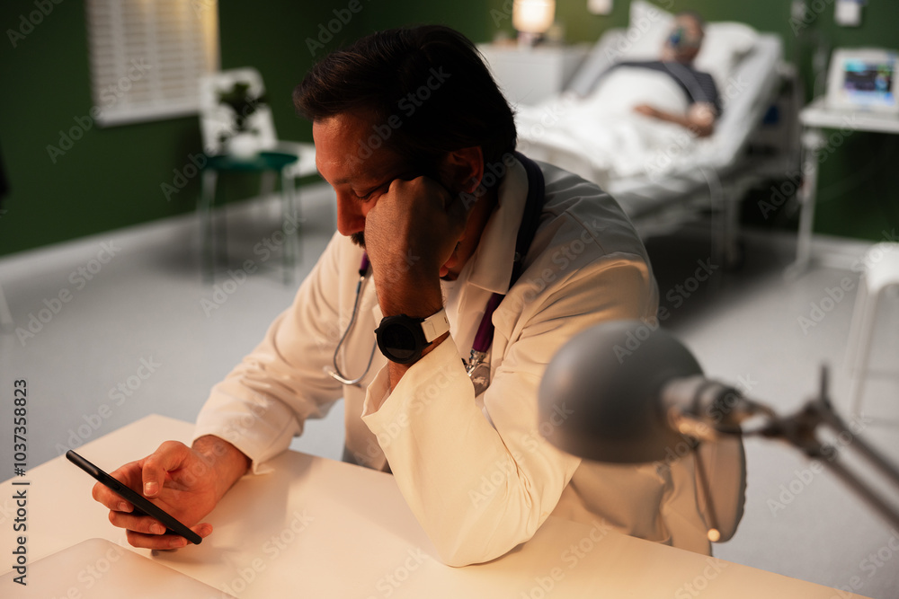 Naklejka premium A middle-aged doctor sits bored with his phone in hand at his desk in a hospital room. An overtired doctor at work