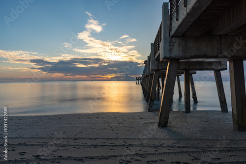 A serene Juno Beach Pier sunrise view under the pier with calm waves and soft reflections, perfect for coastal décor or those seeking tranquil beach imagery with a soothing atmosphere.