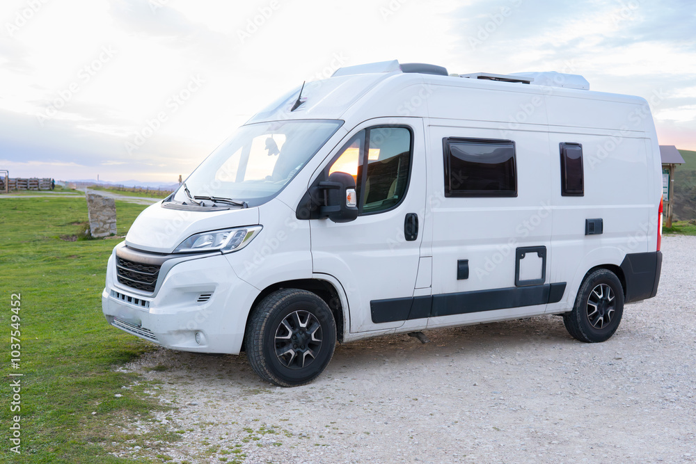 Front and side of a modern white camper van with roof cooler, camped in an empty camper area in the wilderness at sunset. Front and side of a modern white camper van with roof cooler, camped in an empty camper area in the wilderness at sunset.