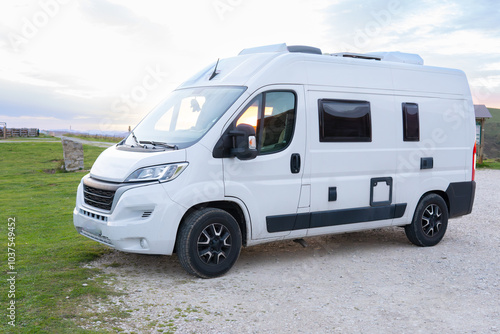 Front and side of a modern white camper van with roof cooler, camped in an empty camper area in the wilderness at sunset.