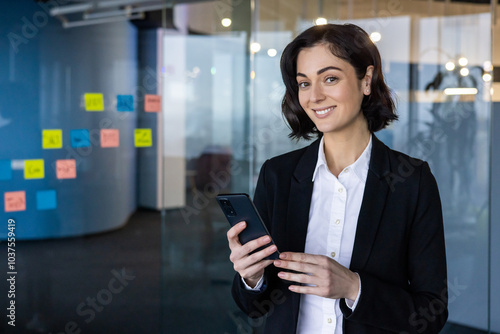 Confident businesswoman using smartphone in office setting, standing near colorful sticky notes on glass wall. Depicts professional technology use, modern collaboration, and workplace strategy.