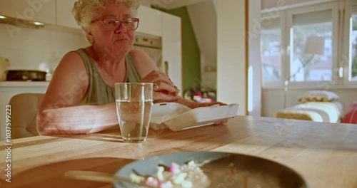 An elderly woman sits alone at her kitchen table eating a takeout meal. She appears contemplative and sad, highlighting the social isolation experienced by many seniors.