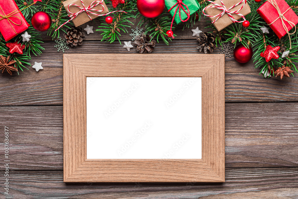 Obraz premium Blank photo frame with Christmas decorations, fir tree branches, gift boxes and pine cones on rustic wooden table. Flat lay. Mock up. Happy New Year and winter holidays layout. Top view