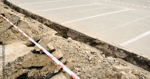 A freshly dug trench along a parking lot, marked with red and white striped safety tape. The scene highlights active construction work and safety measures, showcasing urban development in progress.