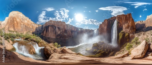 A panoramic view of a powerful waterfall cascading down a canyon in a desert environment. The water crashes into a pool below, creating a mist that hangs in the air.