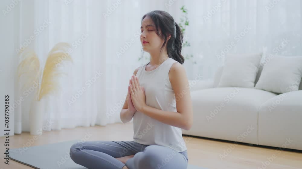Happy young Asian woman practicing yoga and meditation at home sitting on floor in living room in lotus position and relaxing with closed eyes. Mindful meditation and wellbeing concept Happy young Asian woman practicing yoga and meditation at home sitting on floor in living room in lotus position and relaxing with closed eyes. Mindful meditation and wellbeing concept