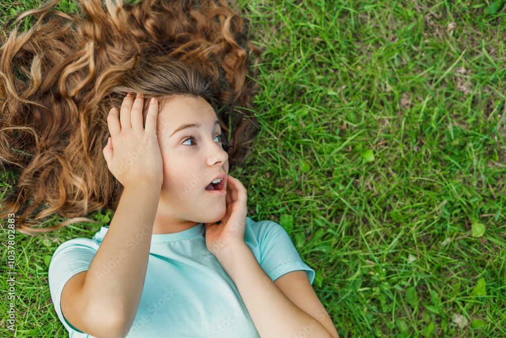 Photo of surprised young girl child in blue t-shirt gasping in amazement, looking with scared and admiring expression to the side, posing against natural green grass background Photo of surprised young girl child in blue t-shirt gasping in amazement, looking with scared and admiring expression to the side, posing against natural green grass background