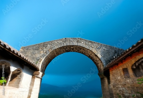 A stone archway stands against a vibrant blue sky, with out of focus ancient stone walls on both sides.