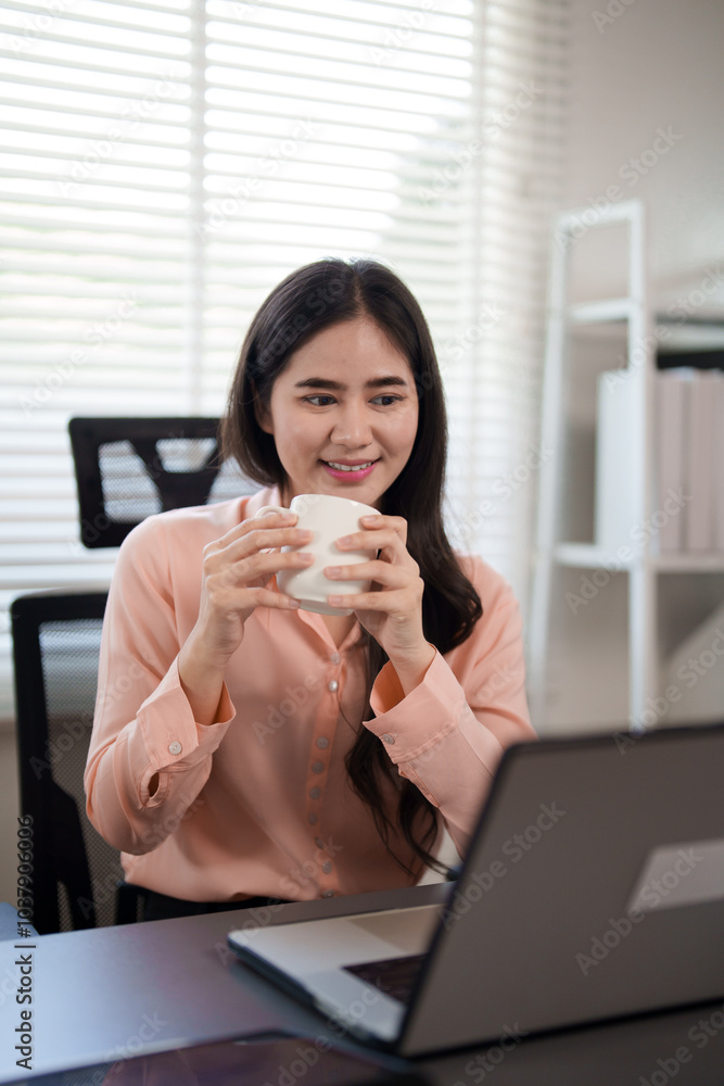 A young woman is sitting at her desk, holding a cup and smiling while looking at her laptop. She is working in a cozy and modern office environment, enjoying a break during her work. A young woman is sitting at her desk, holding a cup and smiling while looking at her laptop. She is working in a cozy and modern office environment, enjoying a break during her work.