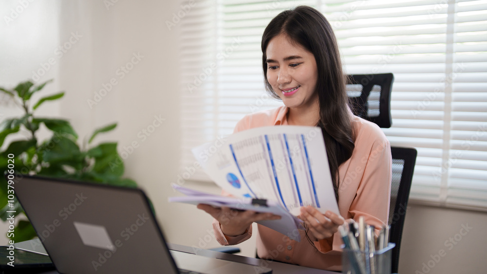 A young woman is sitting at her desk, reviewing documents while working on her laptop in a modern office A young woman is sitting at her desk, reviewing documents while working on her laptop in a modern office