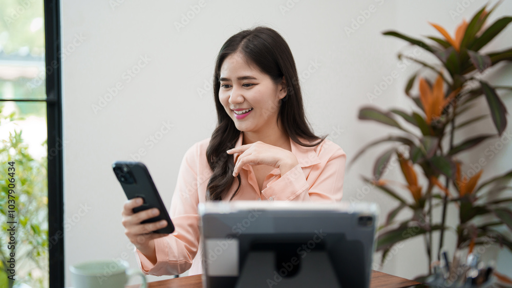 A young woman is sitting at her desk, smiling as she looks at her phone while working on her laptop. She is in a bright and cozy workspace, surrounded by office supplies and greenery A young woman is sitting at her desk, smiling as she looks at her phone while working on her laptop. She is in a bright and cozy workspace, surrounded by office supplies and greenery