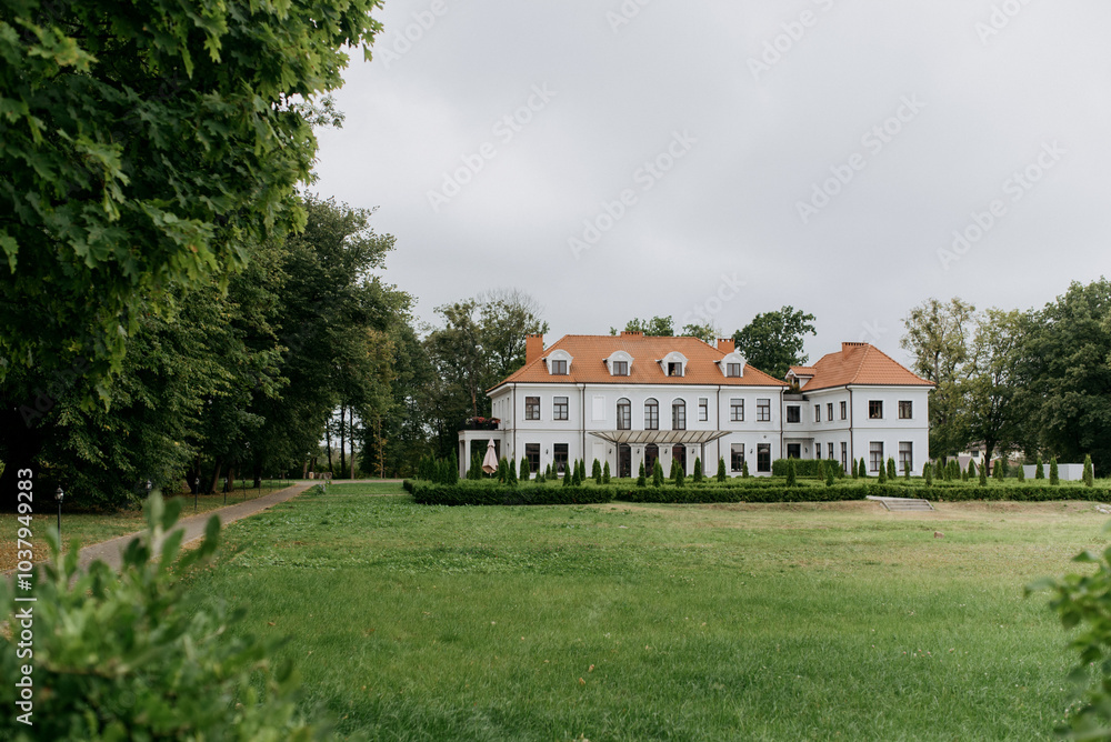 A large, charming white house featuring a striking red roof is beautifully surrounded by lush trees and a vibrant grassy lawn A large, charming white house featuring a striking red roof is beautifully surrounded by lush trees and a vibrant grassy lawn