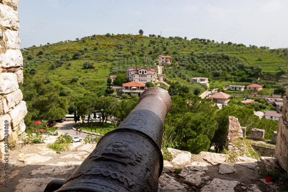 Cannon in ancient stone archway of Berat castle framing picturesque landscape of rolling green hills in Albania, Europe. Historic building of weathered stone and rustic charm. Valley with remote towns Cannon in ancient stone archway of Berat castle framing picturesque landscape of rolling green hills in Albania, Europe. Historic building of weathered stone and rustic charm. Valley with remote towns