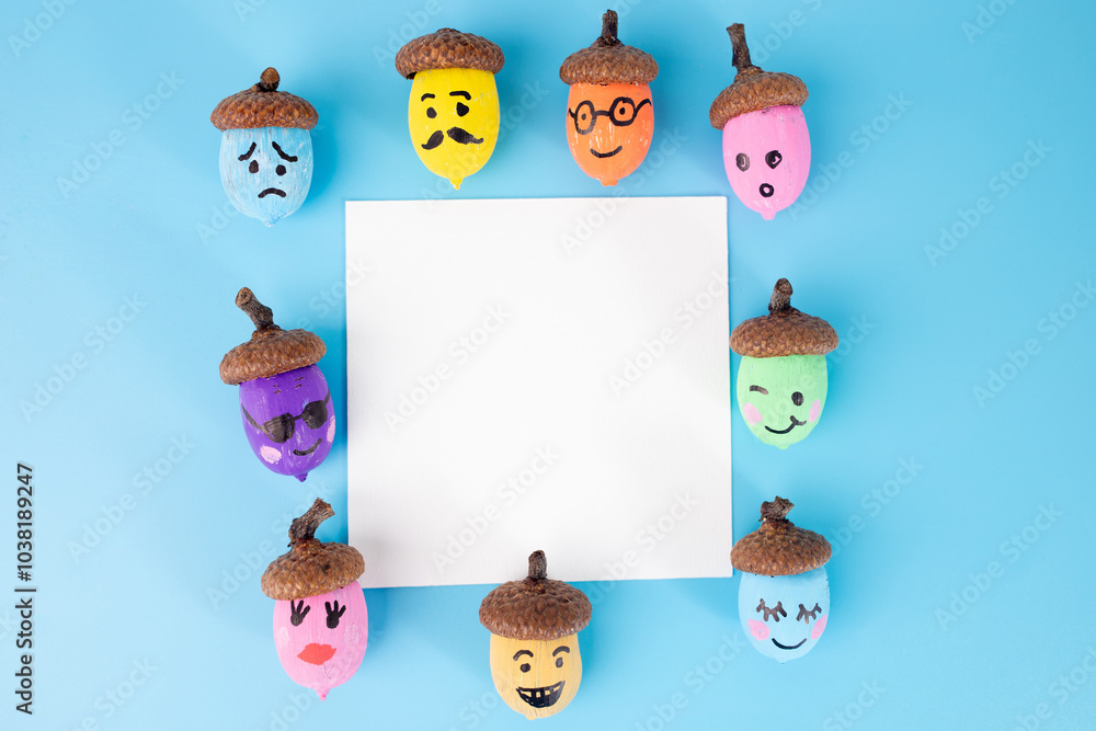 Fototapeta premium Painted emoji acorns, unpainted acorns, brushes, and palette on blue background. Flat lay showcasing creative autumn DIY, perfect for posters, greeting cards, festive designs.