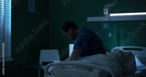 Desperate man seated at edge of bed, trembling, gazes hopelessly out the window, lost in thoughts.