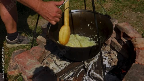 A traditional Romanian cooking scene showing onions being sautéed in a cauldron over a wood fire, a key step in making authentic zacusca. The rustic outdoor setup highlights the time-honored methods. photo