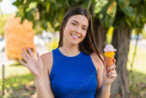 Young pretty Brazilian woman with a cornet ice cream at outdoors saluting with hand with happy expression photo