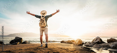 Happy man with backpack standing with arms up at the beach - Delightful tourist enjoying summer vacation by the seaside - Traveling life style and well being concept