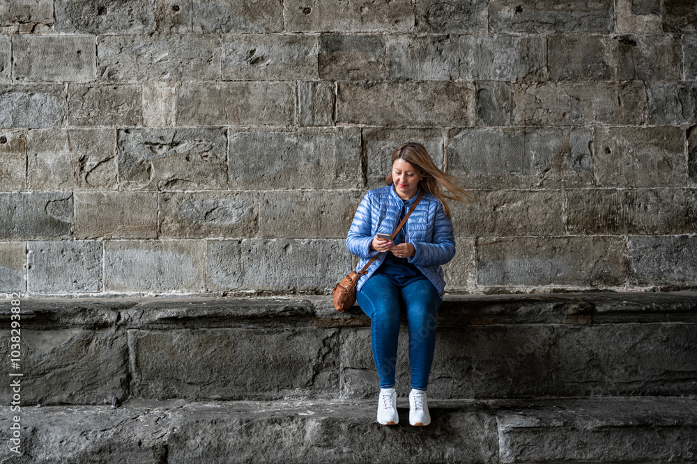 Beautiful young woman wearing jeans and jacket sitting on stone wall against background of old building doing something on smartphone on autumn day. Break in sightseeing in old town. Bergamo, Italy. Beautiful young woman wearing jeans and jacket sitting on stone wall against background of old building doing something on smartphone on autumn day. Break in sightseeing in old town. Bergamo, Italy.