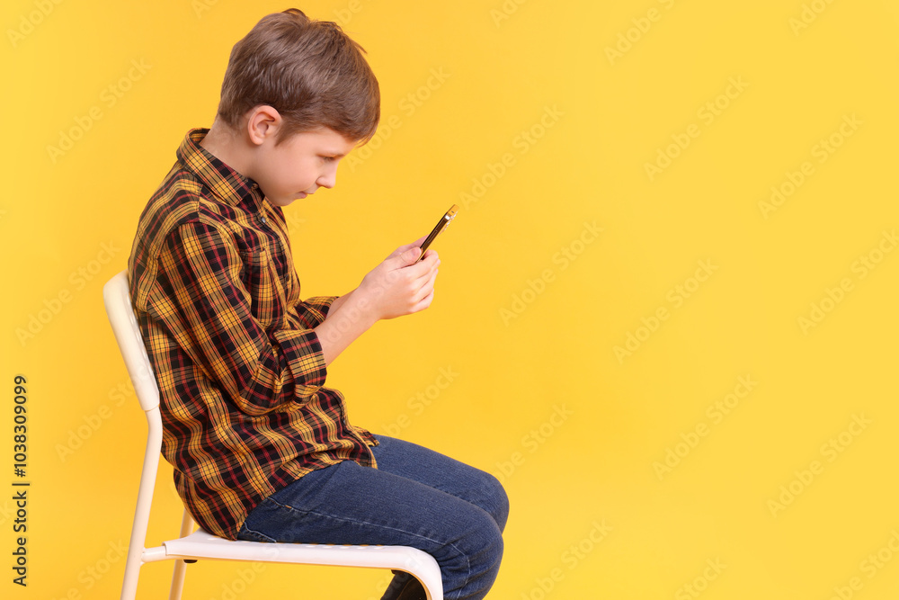 Fototapeta premium Boy with incorrect posture and smartphone sitting on chair against yellow background
