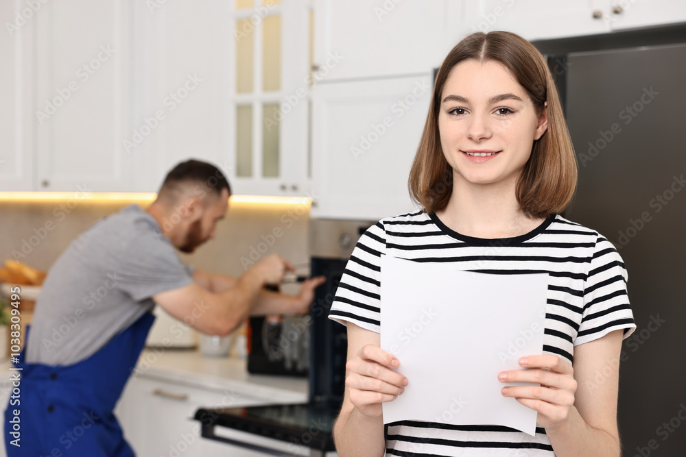 Smiling woman with sheet of paper and repairman fixing oven in kitchen, selective focus Smiling woman with sheet of paper and repairman fixing oven in kitchen, selective focus