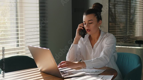 Young busy business woman manager using laptop and mobile cell phone devices in home office. Professional female executive holding smartphone, working on laptop sitting at desk.
