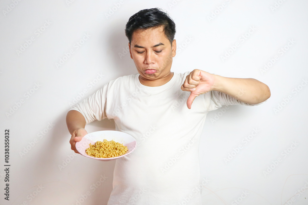 Young asian man with noodle in plate showing thumb down sign Young asian man with noodle in plate showing thumb down sign