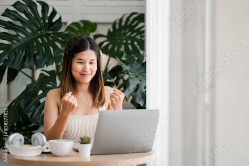 Happy woman enjoying success at home workspace with laptop and tropical plants, expressing excitement and achievement.