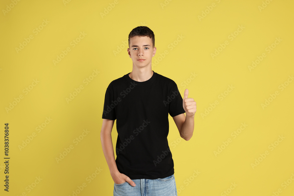 Teenage boy wearing black t-shirt and showing thumbs up on yellow background Teenage boy wearing black t-shirt and showing thumbs up on yellow background