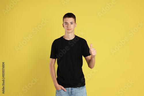 Teenage boy wearing black t-shirt and showing thumbs up on yellow background