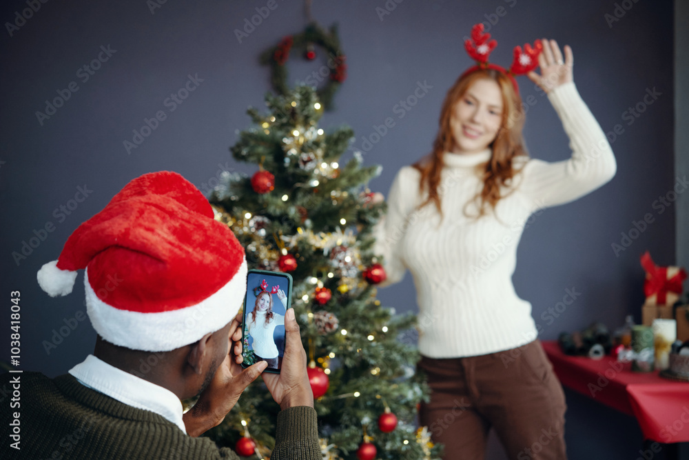 Woman wearing reindeer antlers posing by Christmas tree while man capturing photo with smartphone. Room decorated with festive holiday decorations and ornaments Woman wearing reindeer antlers posing by Christmas tree while man capturing photo with smartphone. Room decorated with festive holiday decorations and ornaments