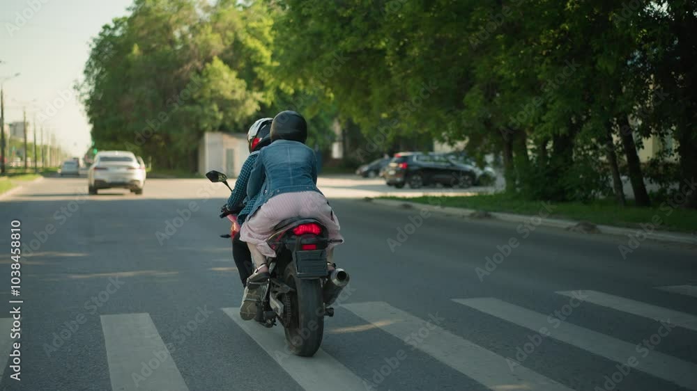 Two women ride a power bike on an urban road. The passenger holds onto the rider as they pass a pedestrian signpost, trees line the road, with an electric pole and a blurred car ahead of them Two women ride a power bike on an urban road. The passenger holds onto the rider as they pass a pedestrian signpost, trees line the road, with an electric pole and a blurred car ahead of them