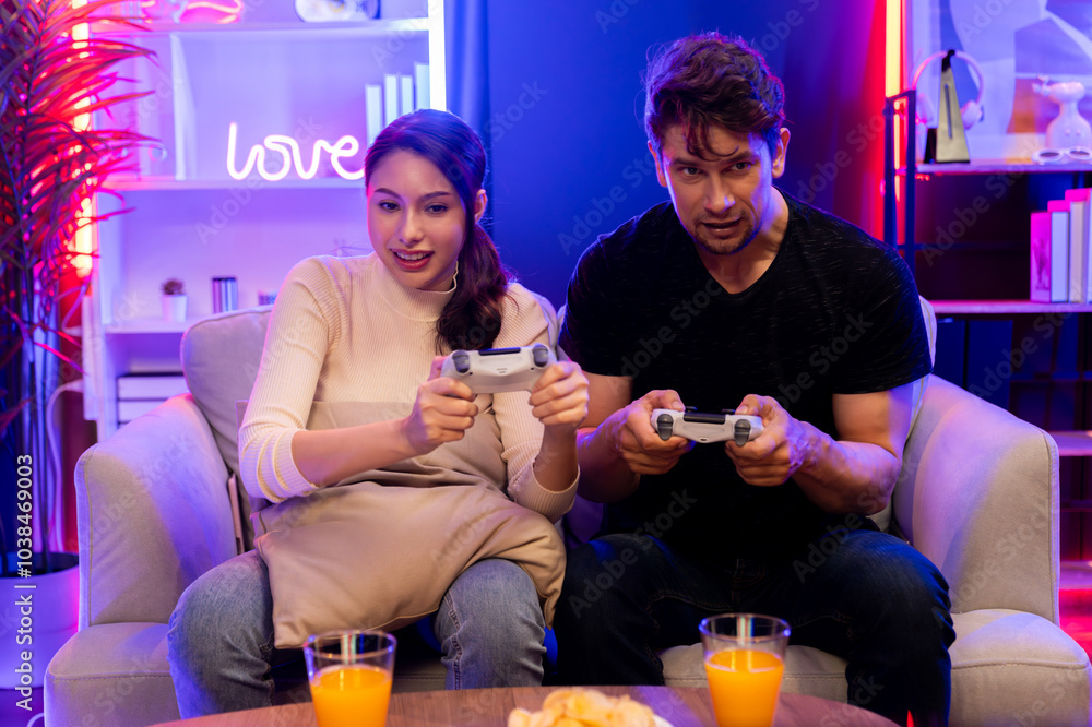 Couple joyful funny players playing video game on TV using joysticks, fighting winner trying to disturb when focusing level challenge in red neon light studio at comfy living home place. Postulate. Couple joyful funny players playing video game on TV using joysticks, fighting winner trying to disturb when focusing level challenge in red neon light studio at comfy living home place. Postulate.