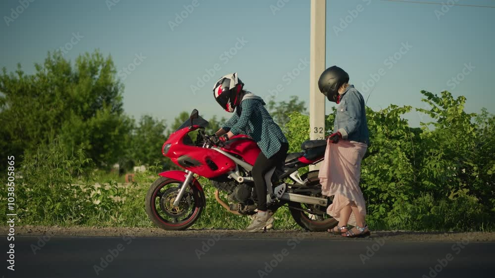 Two women with helmets, one sitting on a red sports bike preparing to move, while the other lady lifts her gown as she climb the bike Two women with helmets, one sitting on a red sports bike preparing to move, while the other lady lifts her gown as she climb the bike