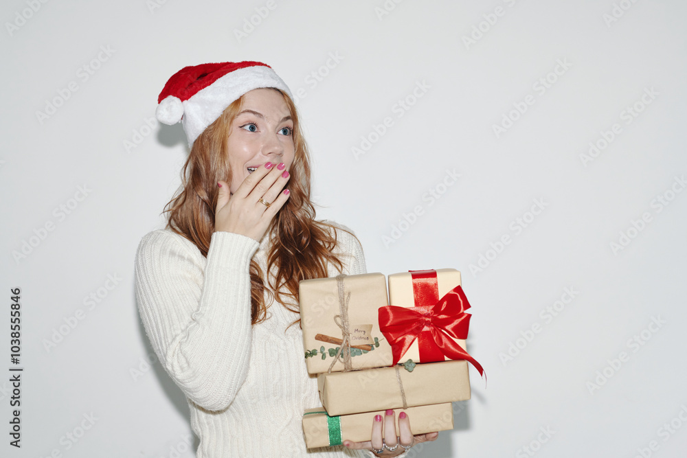 Woman wearing Santa hat looking surprised while holding stack of Christmas gifts wrapped in brown paper and red ribbon. Holiday spirit being illustrated with festive accessories Woman wearing Santa hat looking surprised while holding stack of Christmas gifts wrapped in brown paper and red ribbon. Holiday spirit being illustrated with festive accessories