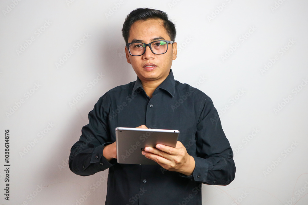 Adult Asian man smiling confident while working using his mobile tablet gadget Adult Asian man smiling confident while working using his mobile tablet gadget