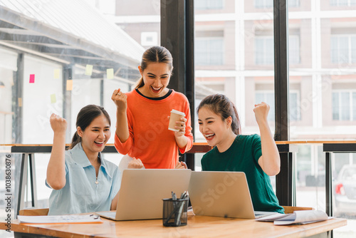 Excited women celebrating success while working on laptops in modern office. Their joyful expressions reflect teamwork and achievement in collaborative environment