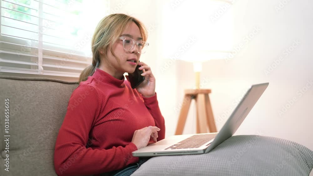 A young woman wearing a red sweater is sitting on a couch, talking on the phone while using a laptop. She looks busy, with a lamp beside her and a cozy home setting A young woman wearing a red sweater is sitting on a couch, talking on the phone while using a laptop. She looks busy, with a lamp beside her and a cozy home setting