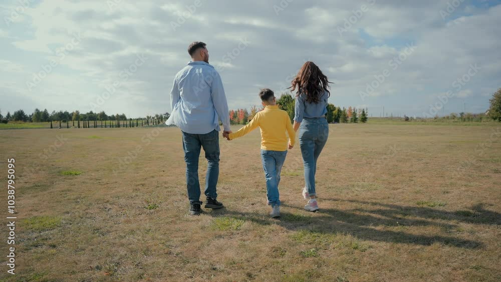Back view Caucasian family stroll talk exploring park field. Mother woman female holding hands father male man hold hand son little boy child kid showing nature talking pointing finger walking outside